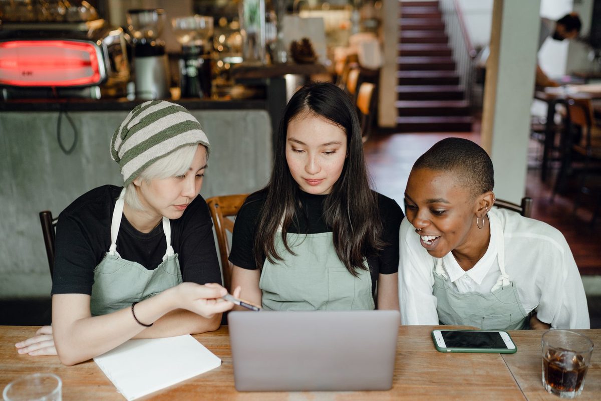 Trois collaboratrices d'un café, en tablier, regardent ensemble un ordinateur portable posé sur une table